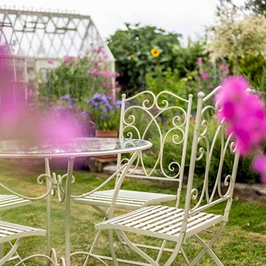 Close-up of cream-coloured Cheltenham 5-piece bistro set on a lawn with flower border and greenhouse in the background.