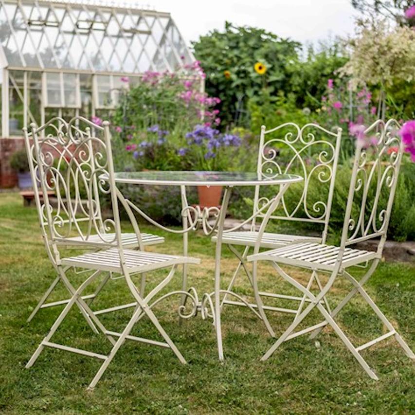 Cream-coloured Cheltenham 5-piece bistro set on a lawn, placed in front of a flower border with a Victorian-style greenhouse in the background.