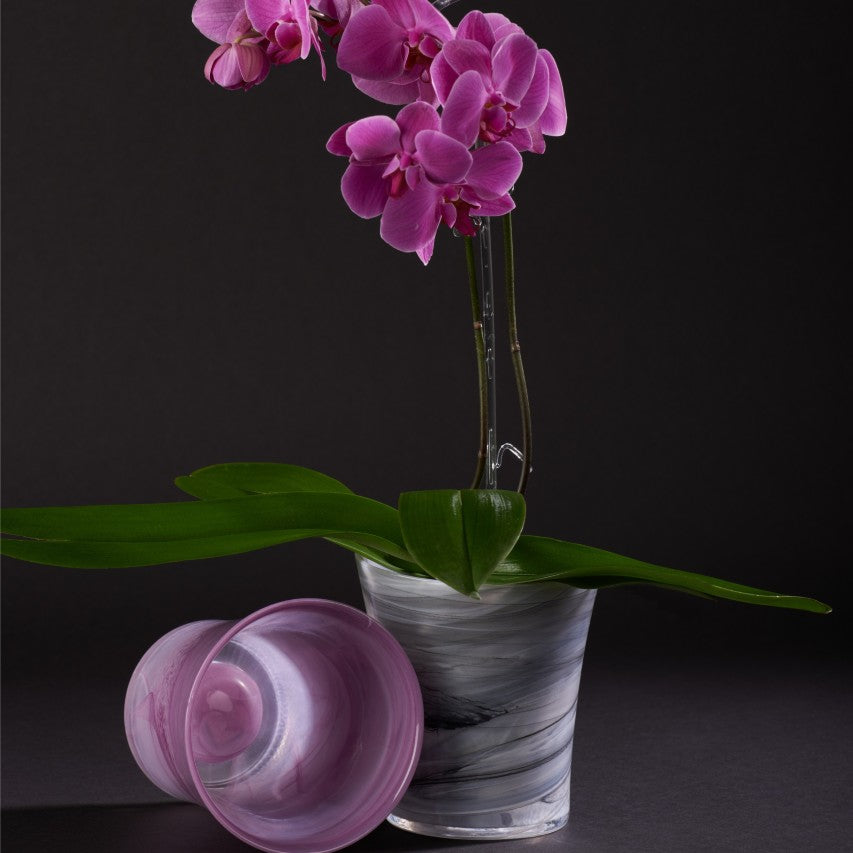 Orchid glass pot in violet on its side next to charcoal orchid glass pot containing pink orchid