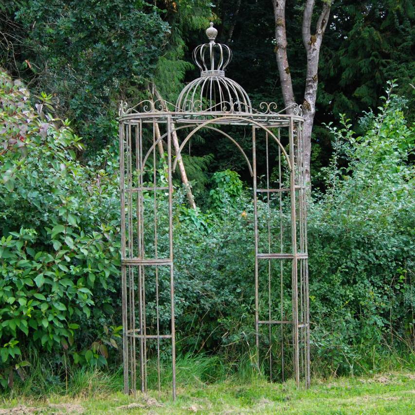 Ascalon ornate archway in antique blue rust standing on grass in front of hedge and trees